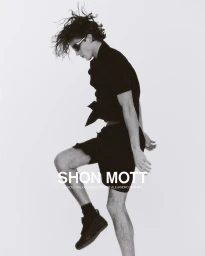 Model in black shorts and shirt wearing sunglasses with wild dark hair mid-leap against a plain white background