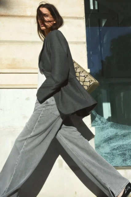 A woman walks along a city street wearing a gray blazer, wide-leg jeans, and black shoes, carrying a snakeskin-print handbag in daylight.