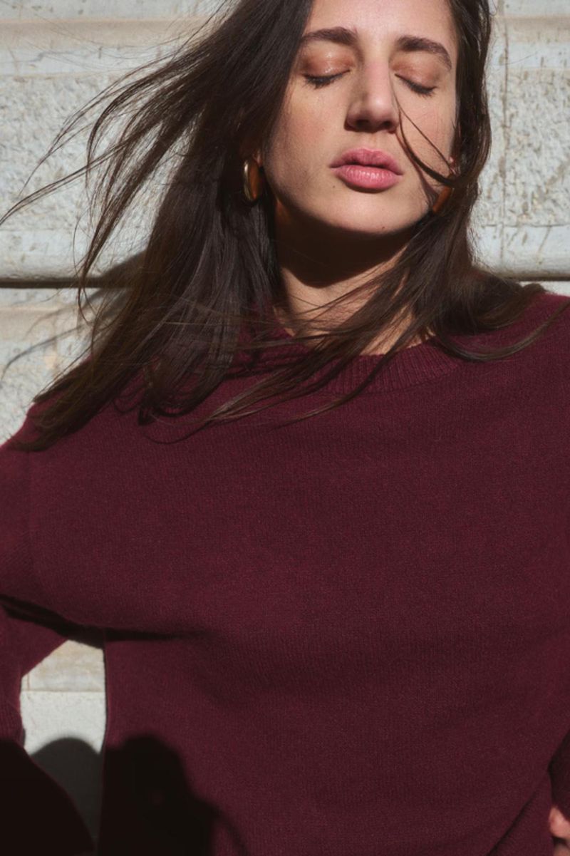 Portrait of a woman with closed eyes and brown hair wearing a maroon sweater in natural outdoor lighting and neutral background.