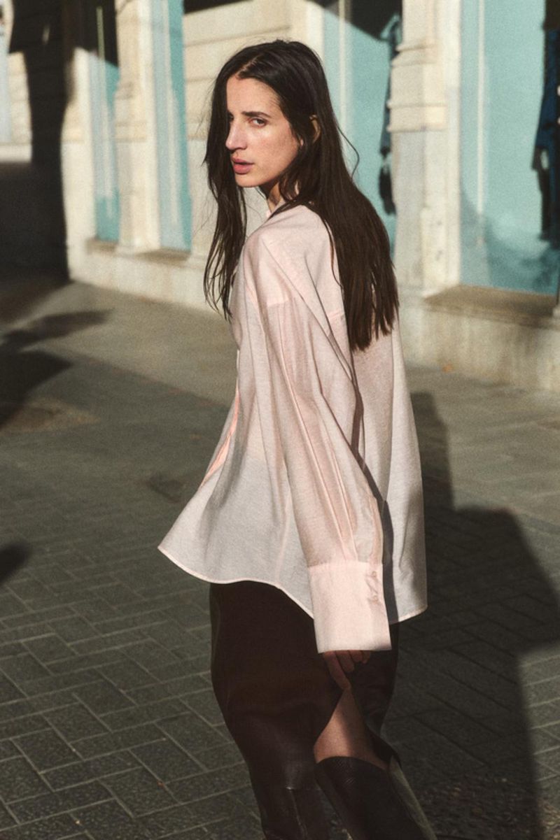 A stylish woman with long dark hair walks on a brick sidewalk wearing a pink button-down shirt, black skirt, and black boots outdoors.