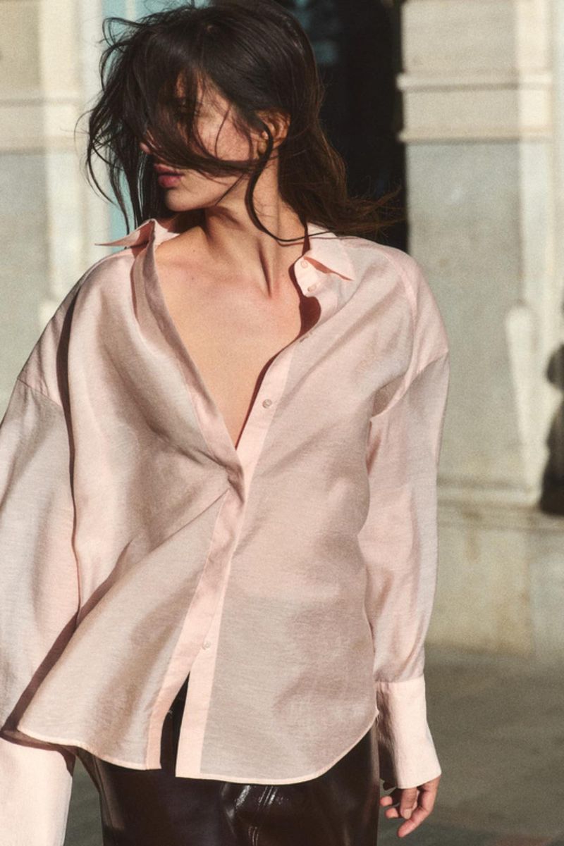 Model wearing a light pink button-down shirt outdoors against a building exterior backdrop with wind-swept hair