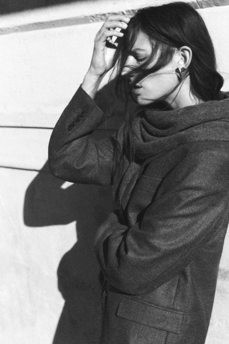 Model in a dark wool coat running hand through wind-blown hair outdoors against a wall casting sharp shadows