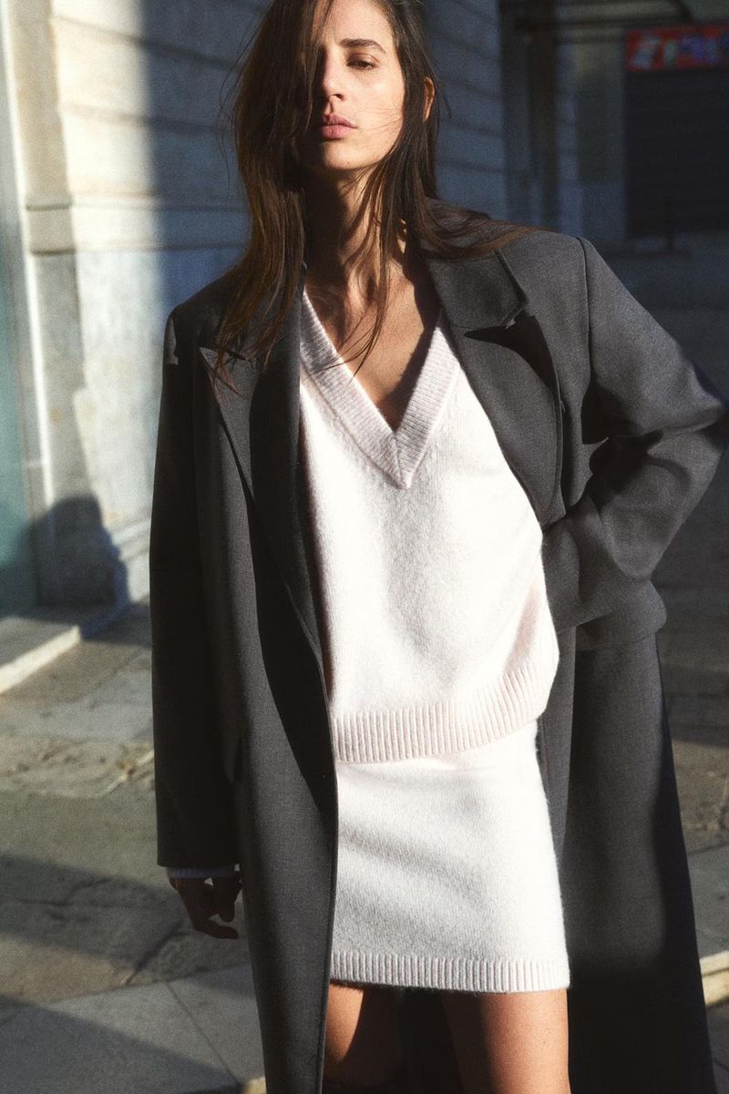 A woman poses outdoors in a knit sweater and matching skirt, styled with an oversized coat. Soft daylight emphasizes the clothing.