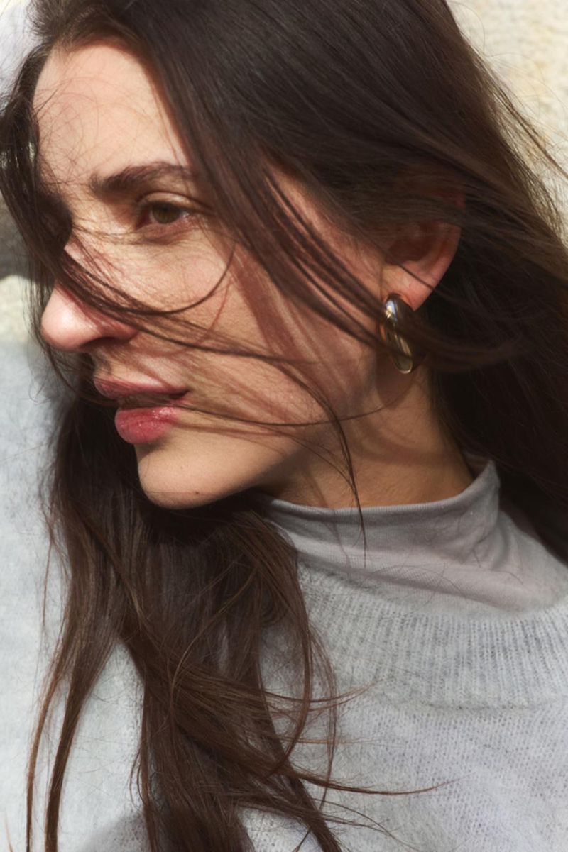 Close-up portrait of a woman with long dark hair blowing across her face, wearing a grey turtleneck and sweater.