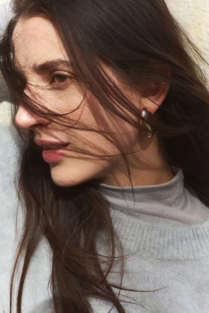 Close-up portrait of a woman with long dark hair blowing across her face, wearing a grey turtleneck and sweater.