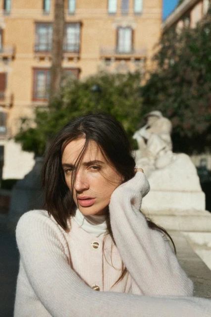 Young woman with long brown hair in a pale pink cardigan poses in front of an urban building and green foliage on a sunny day.