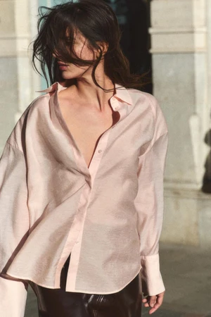 Model wearing a light pink button-down shirt outdoors against a building exterior backdrop with wind-swept hair