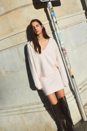 Model in a cream v-neck sweater and matching skirt leaning against a stone building beside a street pole