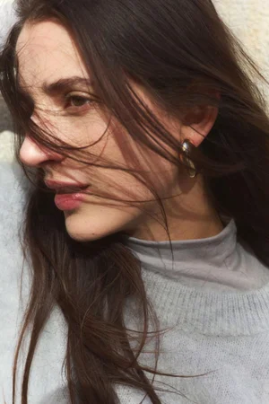 Close-up portrait of a woman with long dark hair blowing across her face, wearing a grey turtleneck and sweater.