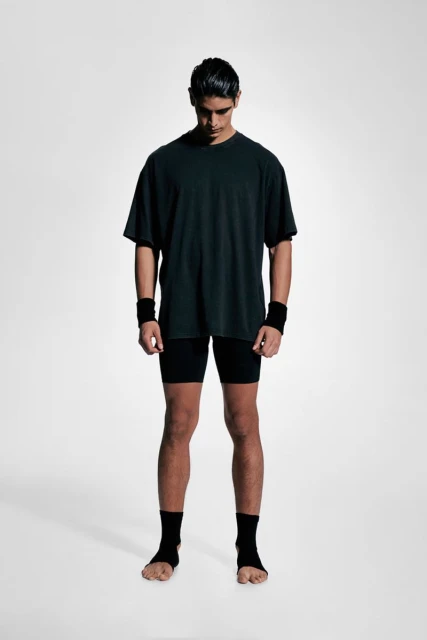 Male athlete stands in photo studio wearing wrist and ankle supports, black t-shirt, and black shorts, looking downward with neutral expression.