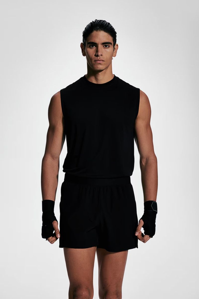 Athletic man poses in black sleeveless shirt and shorts, displaying muscular physique against a bright white studio backdrop.