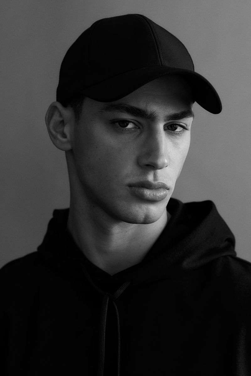 Black-and-white portrait of a young adult male with dark hair, wearing a black cap and hoodie against a neutral gray backdrop.