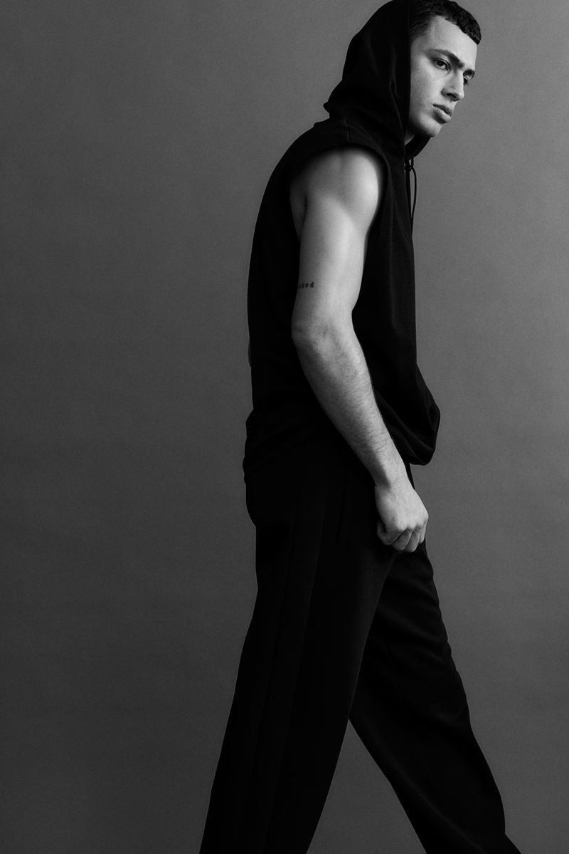 A young adult male is captured in a black-and-white studio portrait, wearing a hooded sleeveless top and pants, shot against a plain gray backdrop.