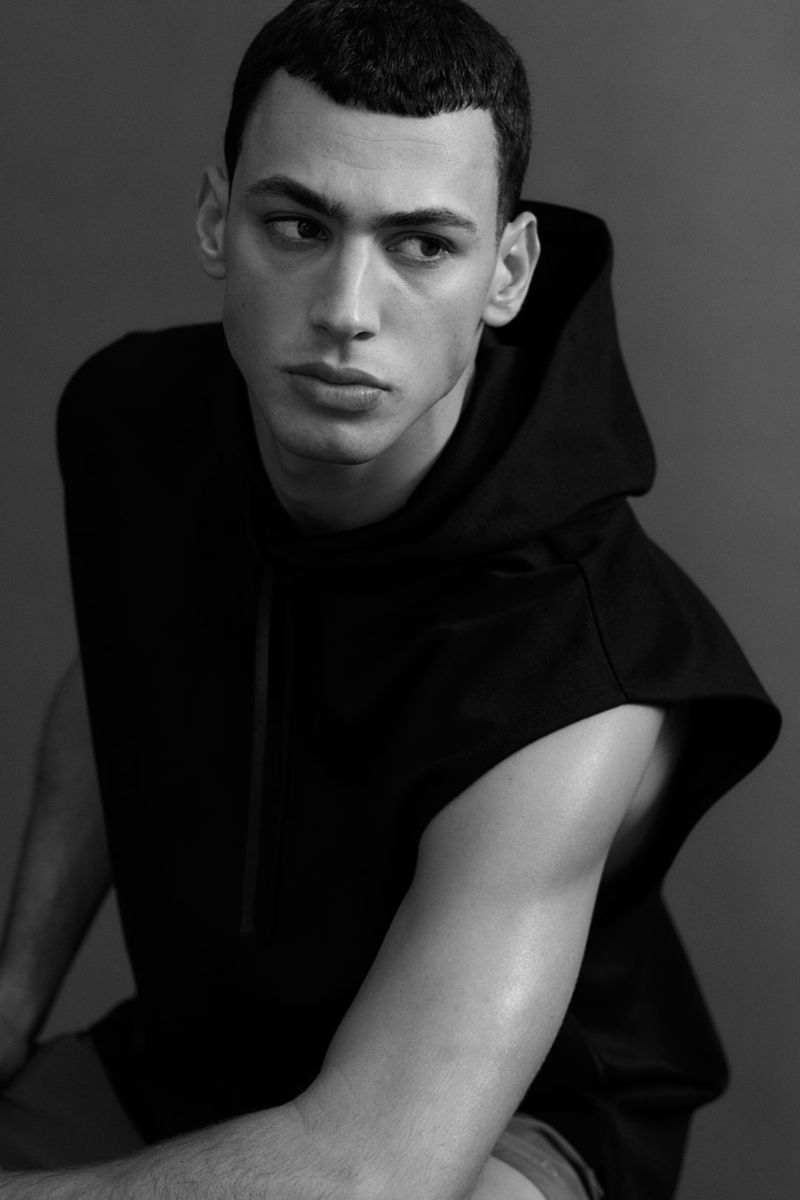 Studio portrait of a young adult man in a black, hooded, sleeveless shirt, looking off to the side against a grey background.