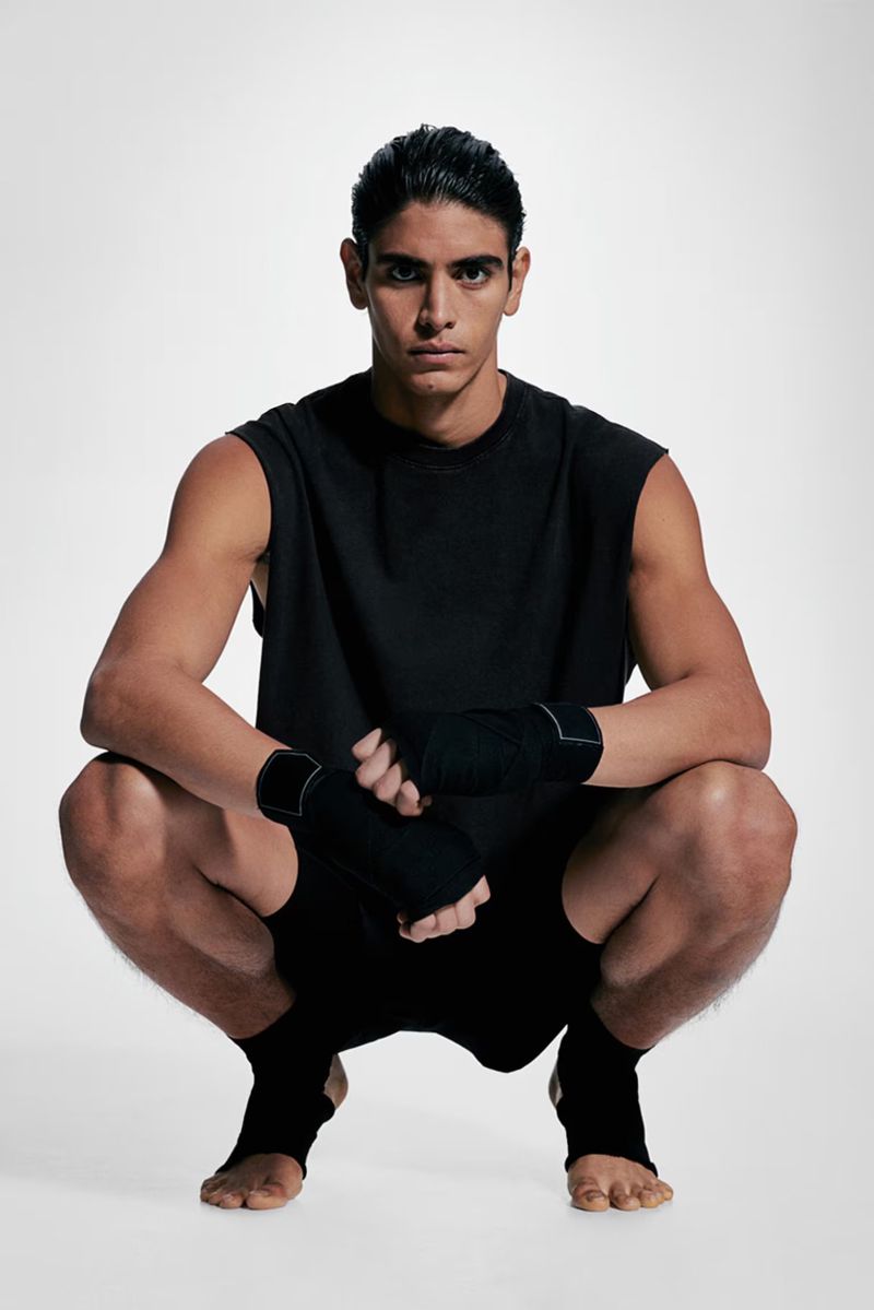 Young man in athletic wear squats in a studio, wearing black top, shorts, hand wraps, and foot wraps. Sports and fitness concept.