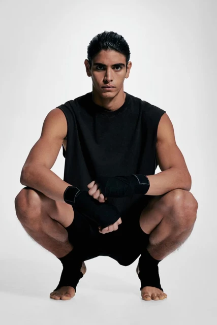 Young man in athletic wear squats in a studio, wearing black top, shorts, hand wraps, and foot wraps. Sports and fitness concept.
