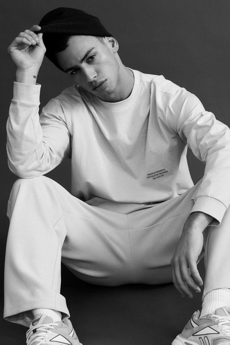 Man wearing a beanie and sweatshirt sitting down against a dark background for the H&M MAN MOVE SS25 campaign.