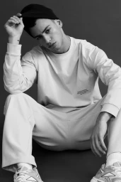 Man wearing a beanie and sweatshirt sitting down against a dark background for the H&M MAN MOVE SS25 campaign.