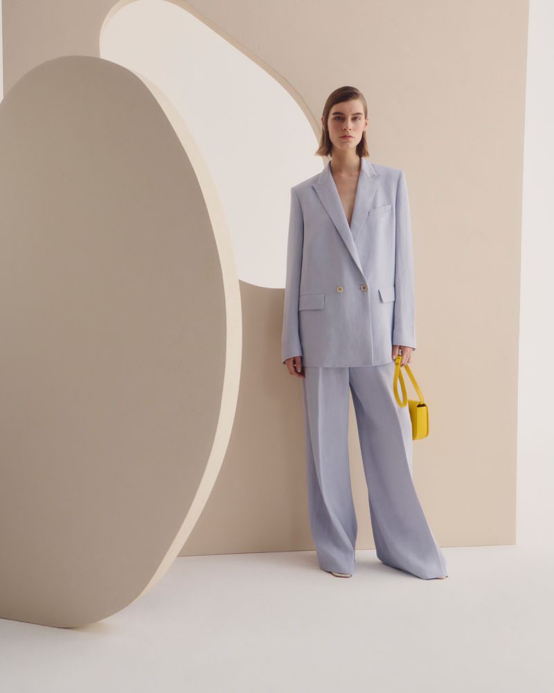 Fashion shot of a woman in a light blue suit holding a yellow purse against a cream-colored backdrop with a circular cutout.