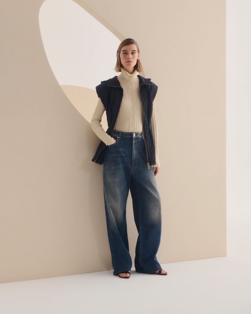 A woman poses in a cream turtleneck, dark blue vest, and wide-leg jeans against a neutral wall with an oval opening.