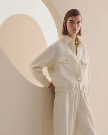 A brunette woman in a relaxed pose wearing a cream-colored shirt and matching pants is featured in a studio setting.