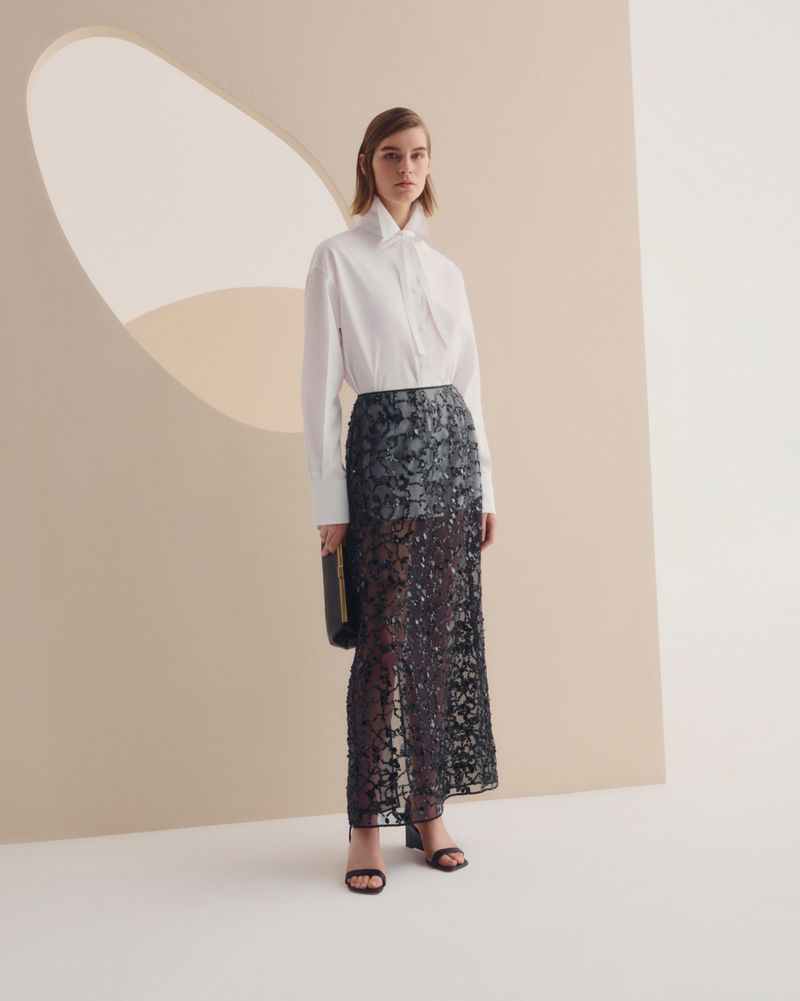 Fashion shoot featuring a woman in a white shirt and black maxi skirt, holding a clutch, posing against a neutral backdrop with a cutout.