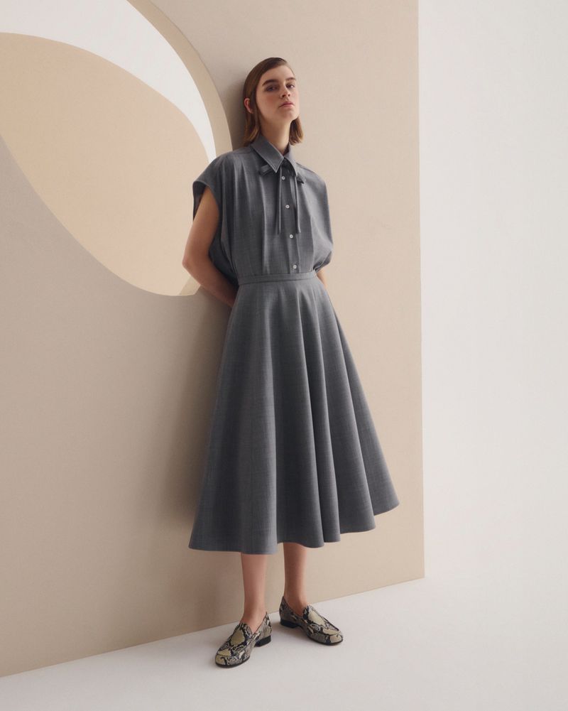 A woman poses in a gray dress with a flared skirt and short sleeves, set against a neutral backdrop with an architectural cutout.