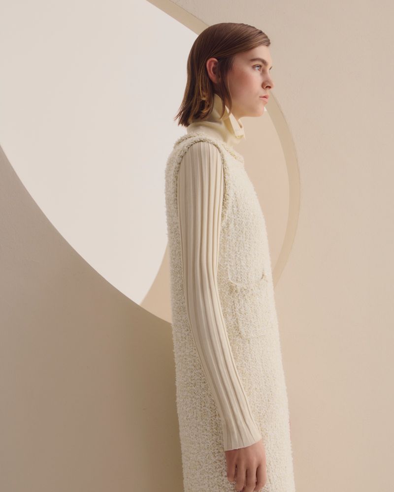 Fashion shot of an adult female model in a sleeveless cream dress and turtleneck top posing in a studio with a light cream background.