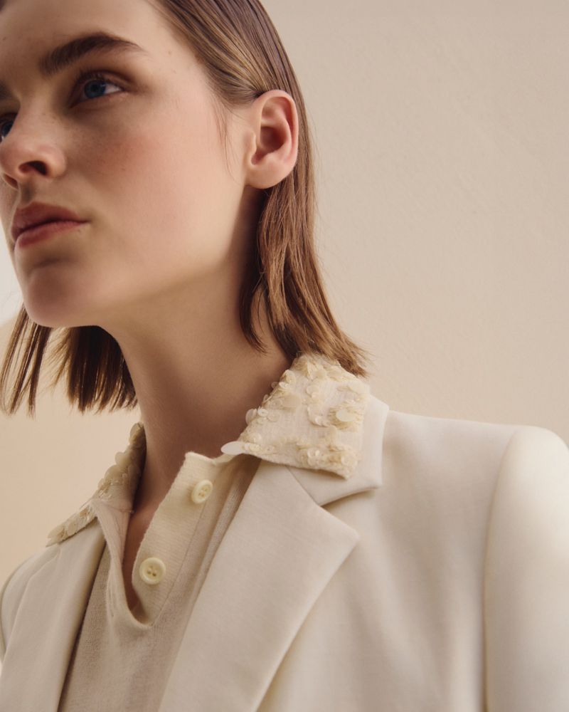 Close up portrait of a female model wearing an off white blazer and matching polo shirt against a plain background.