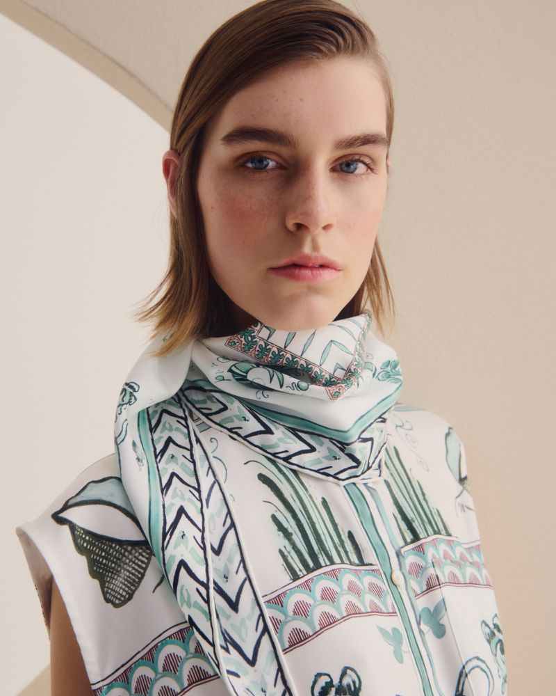 Portrait of a young woman with fair skin and blue eyes wearing a patterned silk scarf, set against a soft, neutral background.