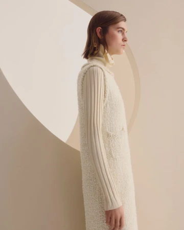 Model in profile wearing a cream sleeveless knit dress over a ribbed turtleneck against a minimalist geometric backdrop