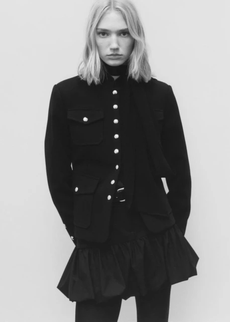 Monochrome portrait of a fair-skinned fashion model with blonde hair wearing a military-style jacket and black miniskirt against a white backdrop.