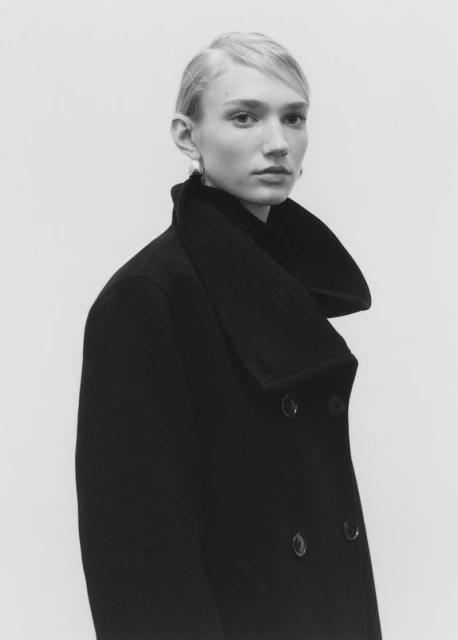 Blonde woman in black coat posing in photo studio setting. Shoulder-length shot with neutral facial expression, minimalistic fashion style.