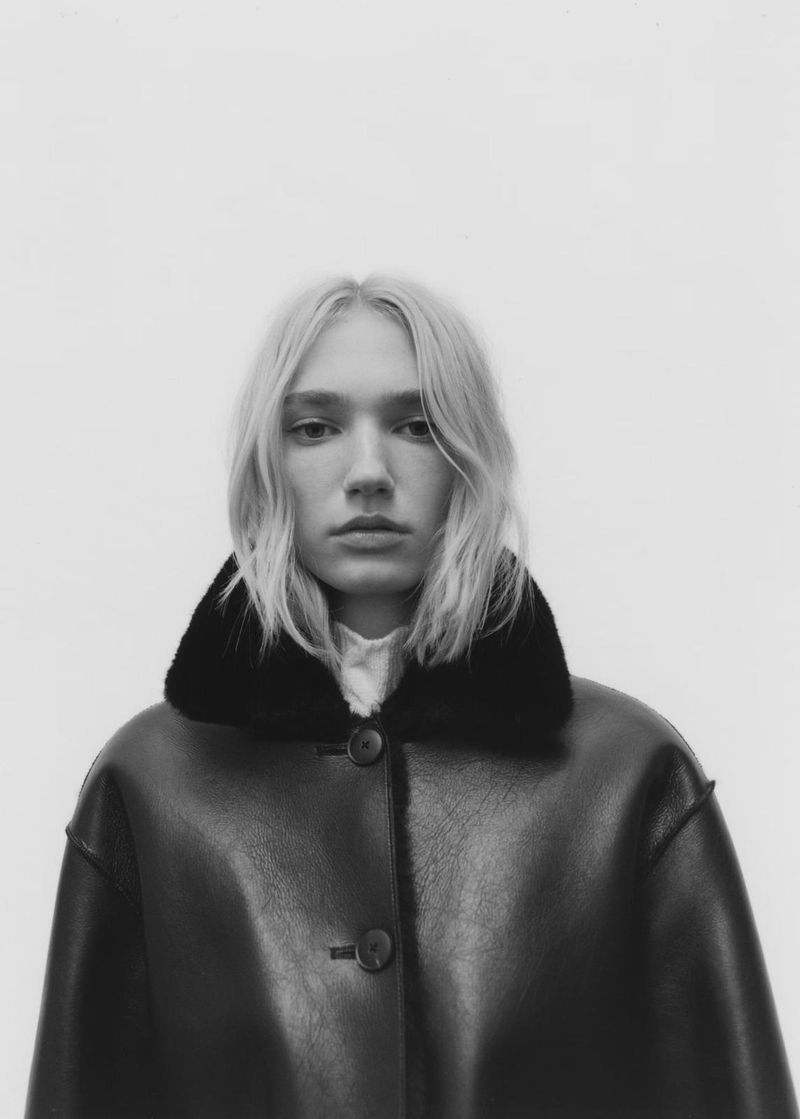 Portrait of a blonde woman wearing a dark jacket with a fur collar against a light background for Mango AW 25.