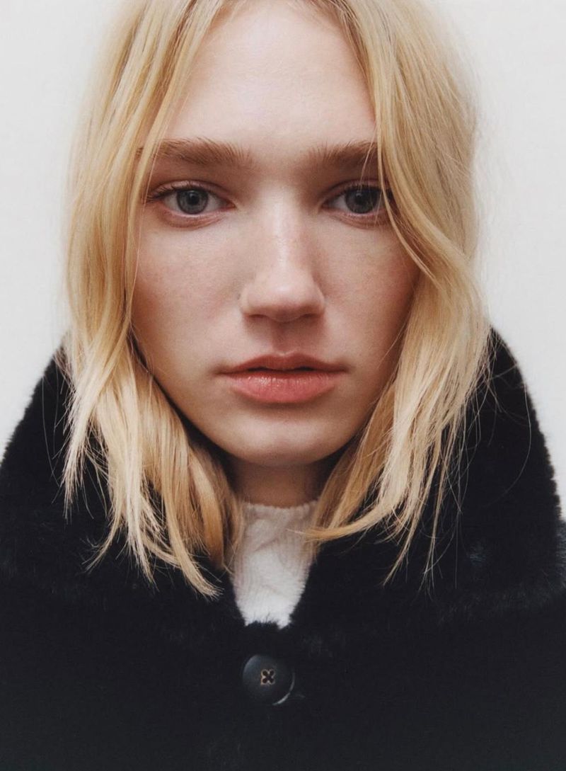 Close-up portrait of a blonde model wearing a black faux-fur collared coat against a plain white studio background.