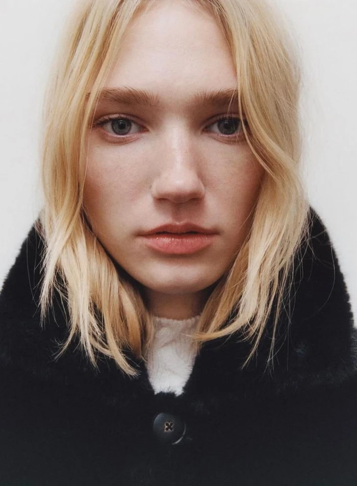 Close-up portrait of a blonde model wearing a black faux-fur collared coat against a plain white studio background.