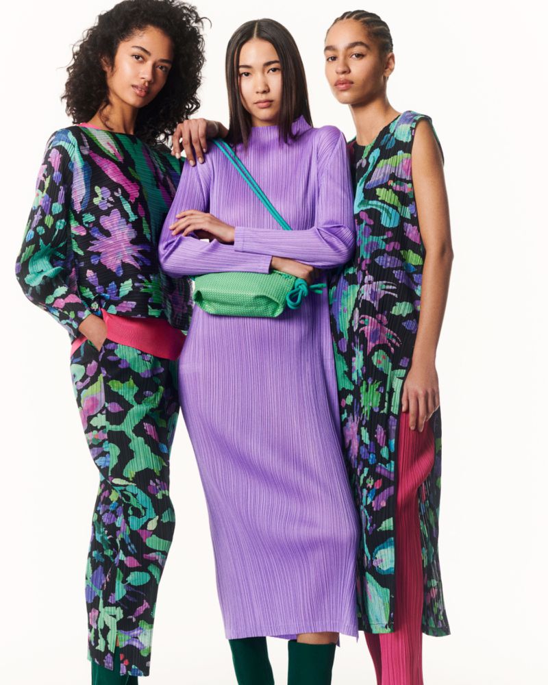 Three fashion models pose in a studio. The models are wearing pleated outfits with a bright, colorful floral pattern and solid colors.
