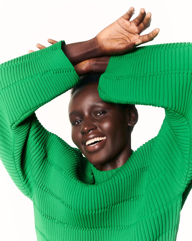 Smiling dark-skinned woman with green ribbed sweater, hands above her head, medium shot, white backdrop. Beauty and fashion concept.