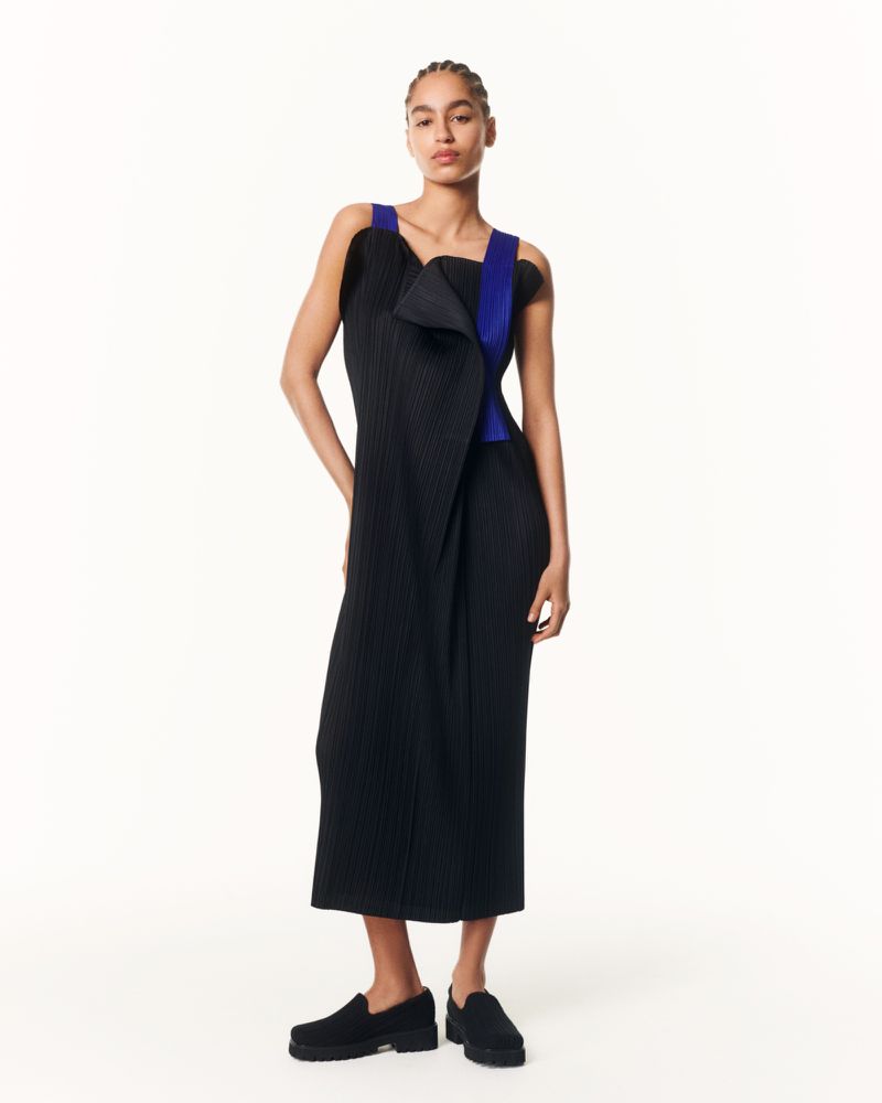 A single female model stands in a bright studio wearing a black dress with royal blue straps and black slip-on shoes, looking at the camera.