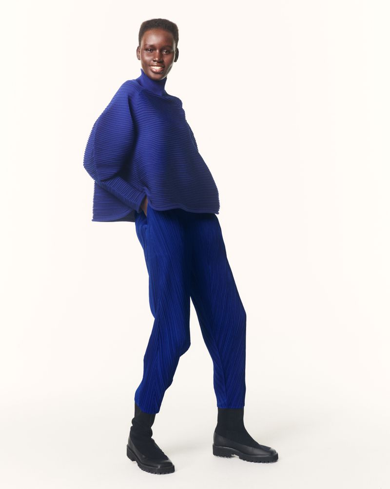 An adult female fashion model poses in a photo studio setting wearing a pleated blue top, pants, and black boots against an off-white backdrop.