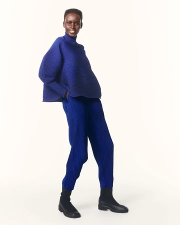 An adult female fashion model poses in a photo studio setting wearing a pleated blue top, pants, and black boots against an off-white backdrop.