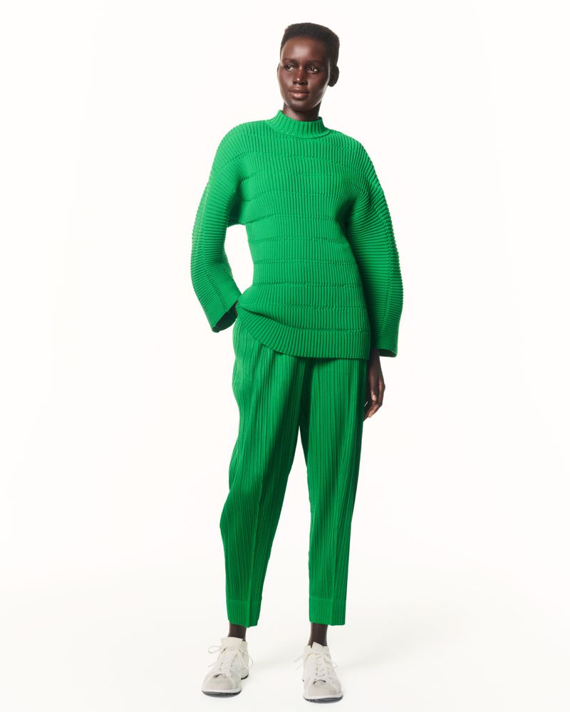 Studio shot of a slender, dark-skinned woman modeling a green ribbed knit sweater and pleated pants, paired with gray sneakers.