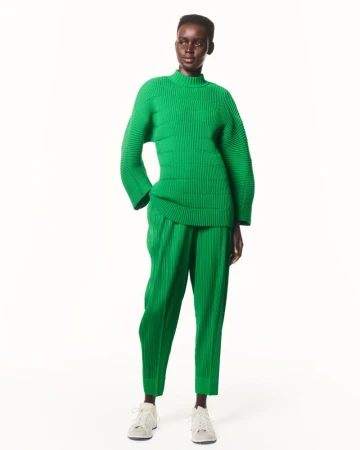Studio shot of a slender, dark-skinned woman modeling a green ribbed knit sweater and pleated pants, paired with gray sneakers.