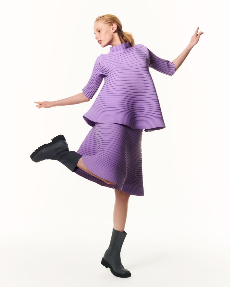 A fair-skinned woman in a lilac knit skirt and top strikes a dynamic pose in a studio setting, emphasizing fashion and style.