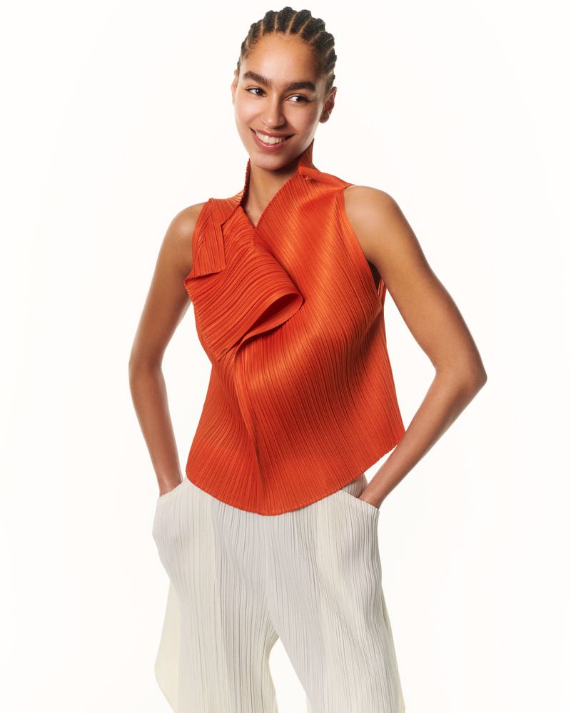 Portrait of a smiling adult woman in a pleated orange sleeveless top and white pants, posed in front of a bright white background.