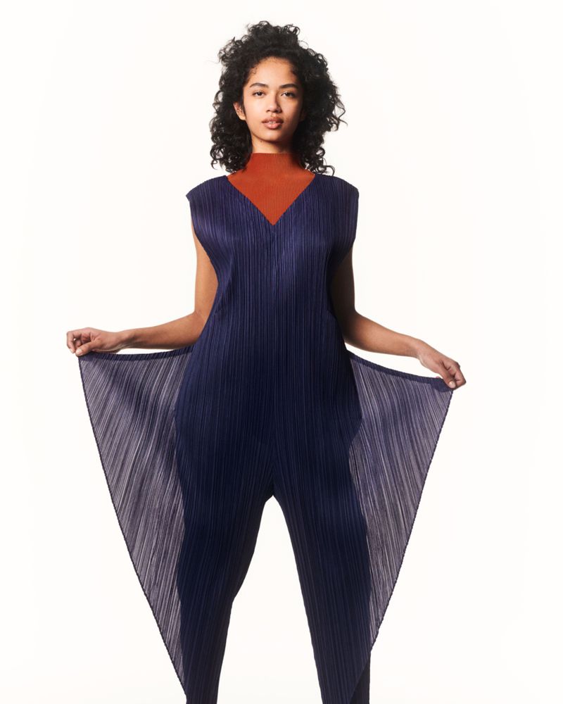 Full-length studio shot of an adult woman posing. She wears a navy pleated jumpsuit over a rust-colored top against a white backdrop.