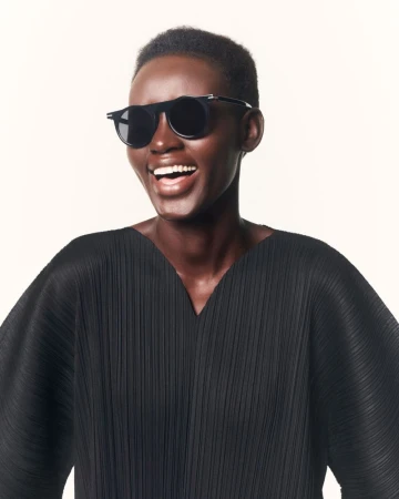A dark-skinned woman smiles, wearing black sunglasses and a pleated top against a light background, showcasing a cheerful fashion portrait.
