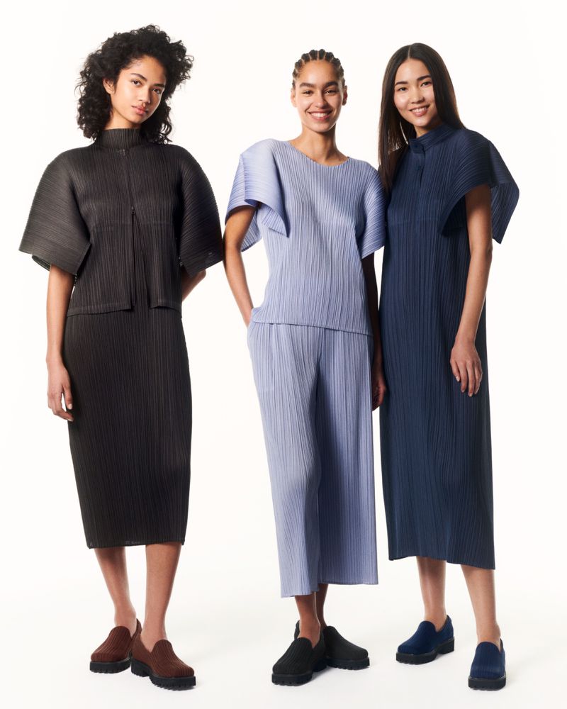 Three fashion models pose in a studio wearing pleated two-piece outfits and matching loafers in black, blue, and periwinkle colors.