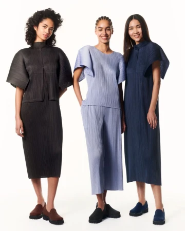 Three fashion models pose in a studio wearing pleated two-piece outfits and matching loafers in black, blue, and periwinkle colors.