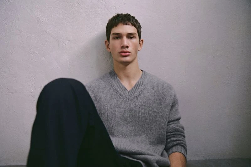 A young adult male poses in a grey v-neck sweater and navy trousers against a textured grey wall in a studio setting.